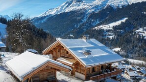 A wooden cabin with a snow-covered roof, surrounded by a snowy landscape and mountains in the background.