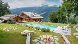 A wooden cabin with a swimming pool, surrounded by a stone pathway and a fire pit area.