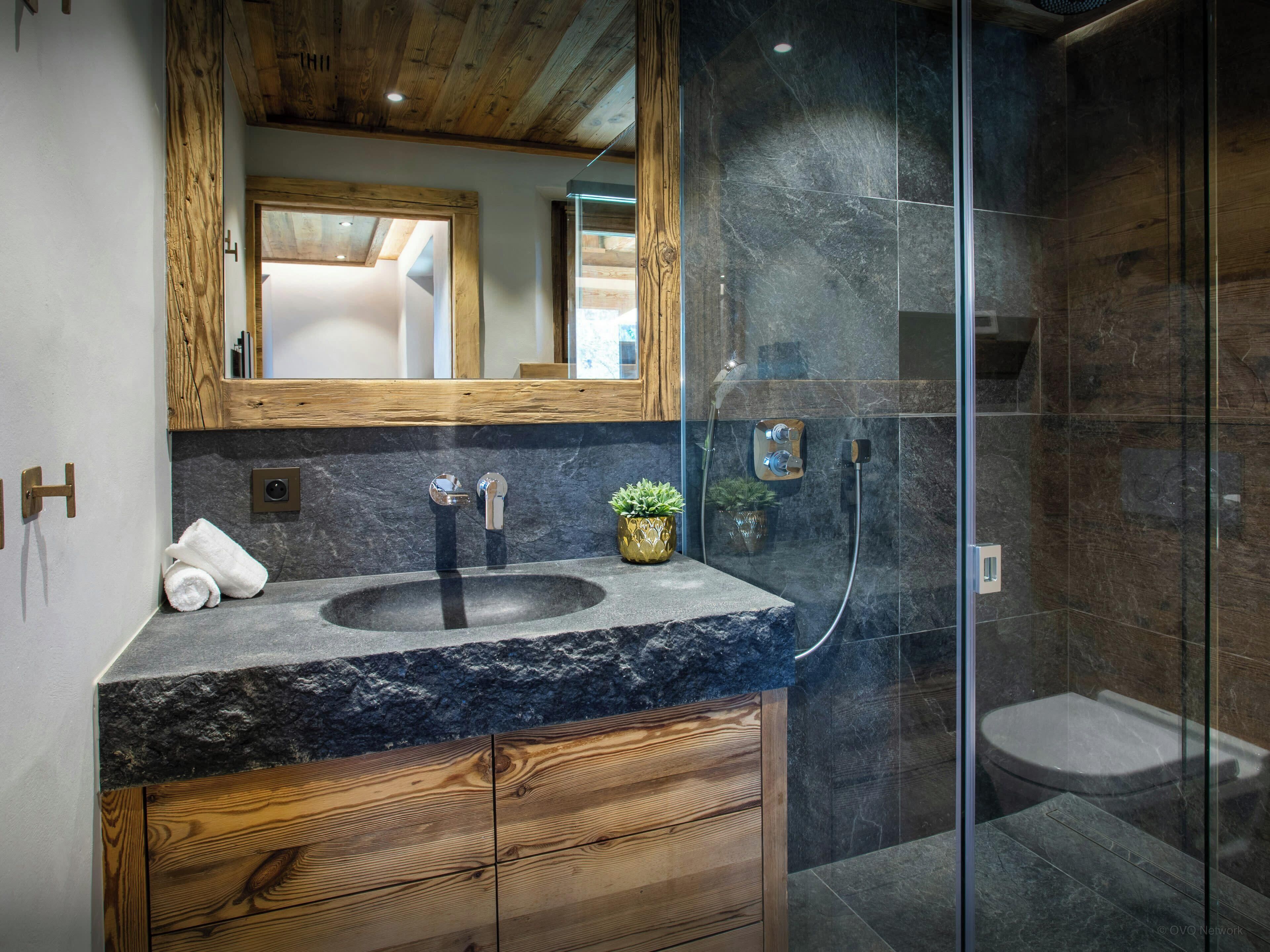Un bagno moderno con un piano di lavoro in granito, mobili in legno, un ampio specchio e una cabina doccia con pareti in vetro.