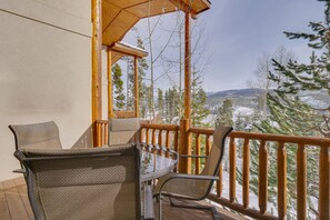 Une terrasse en bois, dotée de meubles d’extérieur et d’une rambarde en bois, offrant une vue sur des arbres et des montagnes enneigés.