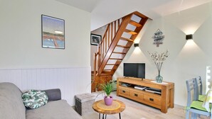 A living room with a wooden staircase, a sofa, a TV on a wooden stand, and a small round table with a plant. - La Maison de Louise - Ferel (FEREL)