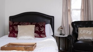 A neatly made bed with a wooden tray, a black leather armchair with a 'Cozy' pillow, and a small table with a lamp and a pair of glasses. - Cozy Parkview Oasis With Gameroom (Amarillo)