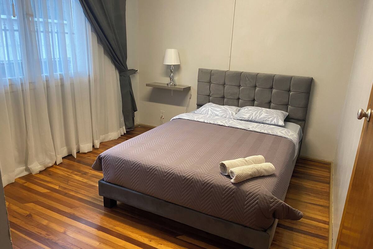 A neatly made bed with a grey headboard, a bedside table with a lamp, and a window with sheer curtains.