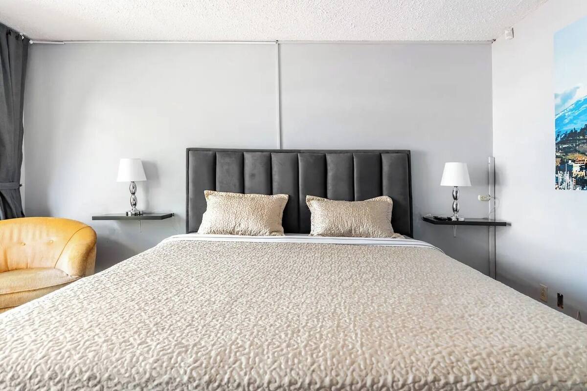 A modern hotel room with a large bed, two bedside lamps, a chair, and a cityscape painting on the wall.