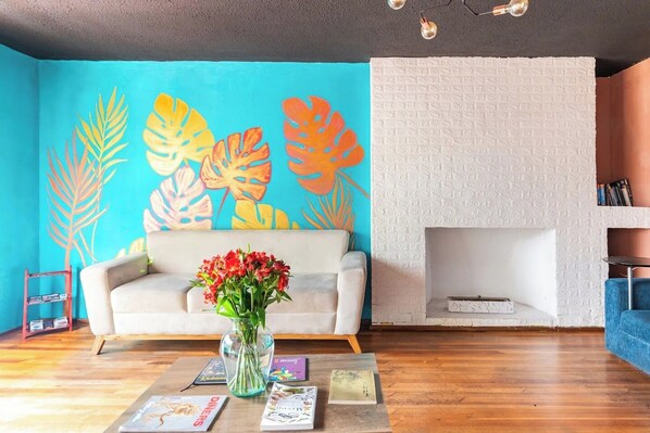 Un salon avec un canapé blanc, une table basse ornée de livres et d’un vase de fleurs, une fresque murale aux couleurs vives et une cheminée en briques blanches.
