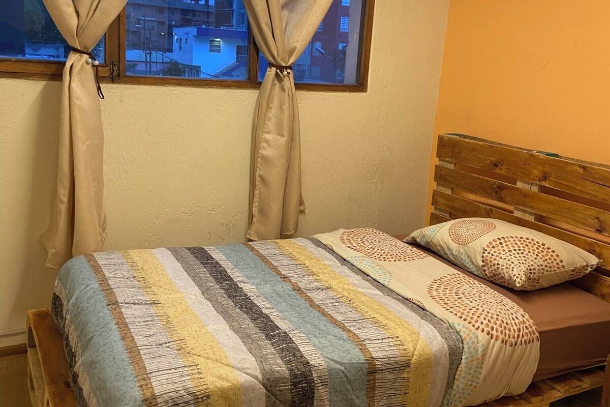 A single bed with a striped blanket and two pillows, a wooden headboard, and a window with curtains tied back.