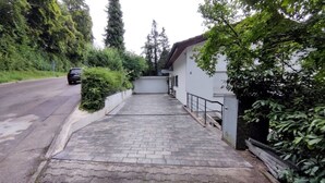 A paved driveway leading to a white building with a grey roof, surrounded by greenery and a parked car on the adjacent road. - Vacation apartment 94 sqm & 16 sqm terrace - with magnificent panoramic views of the city and the Alps (Waldshut-Tiengen)