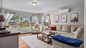 Ein geräumiges Wohnzimmer mit Sofa, Sessel und Couchtisch, großen Fenstern mit Vorhängen und Blick auf einen Garten.