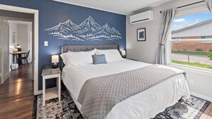 Ein Schlafzimmer mit Bett, Nachttisch, Wandgemälde, Fenster mit Vorhängen und Blick ins Freie.