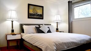 A neatly made bed with white bedding, two bedside tables with lamps, a framed painting of a ship on the wall, and a window with a view of trees. - Cellar House NOTL - Entire Home in the Heart of Niagara-on-the-Lake (Niagara-on-the-Lake)