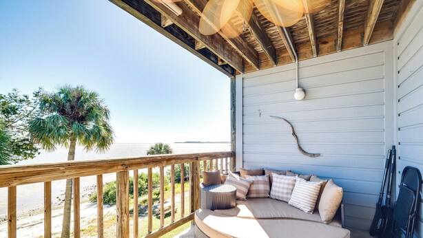 A covered outdoor seating area with a curved sofa and cushions, overlooking a beach and palm trees. - Cedar Key Condo w/ Balcony & Gulf Views! (Cedar Key)