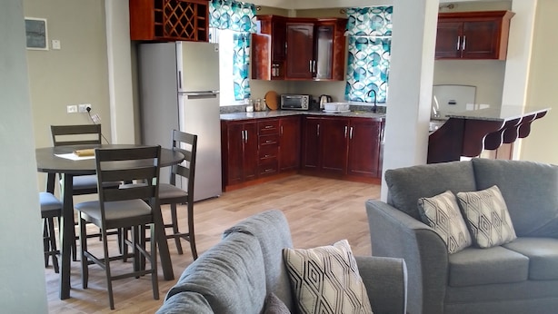 A modern living room with a grey sofa and armchair, a dining area with wooden chairs, and a kitchen with wooden cabinets and a tiled backsplash. - Two Bedroom Apartment Rental (Paix Bouche)