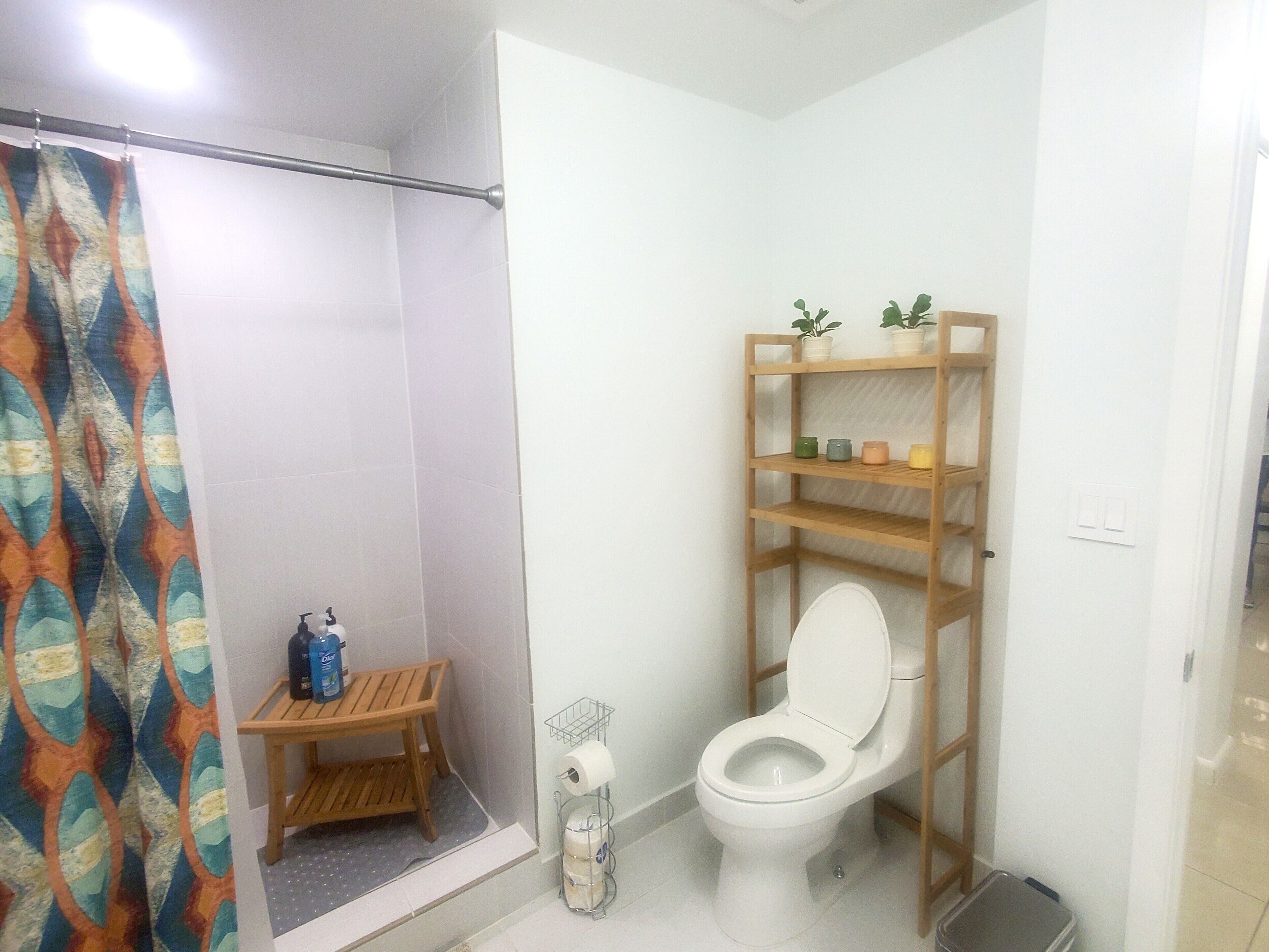 A bathroom with a shower, toilet, shelf with toiletries, and a small wooden stool.
