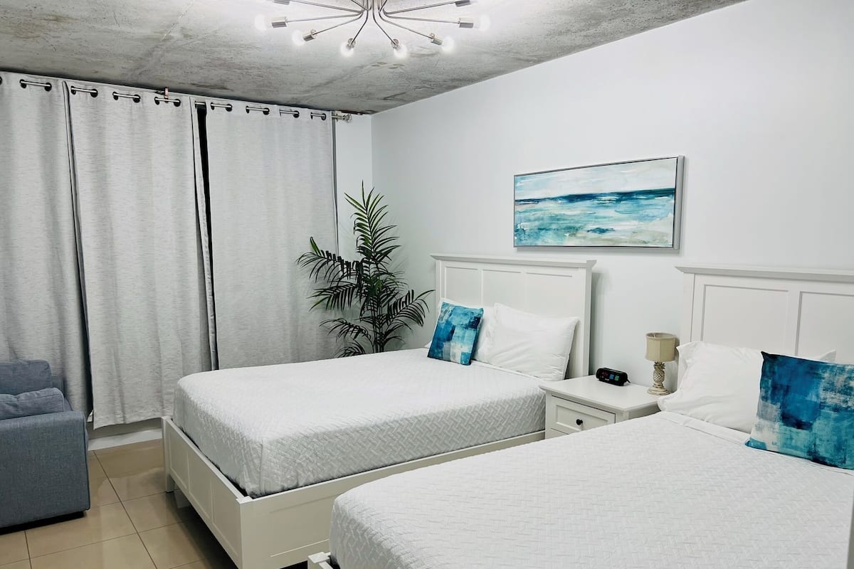 A hotel room with two beds, a grey sofa, a potted plant, a nightstand with a lamp, and a painting of the sea on the wall.