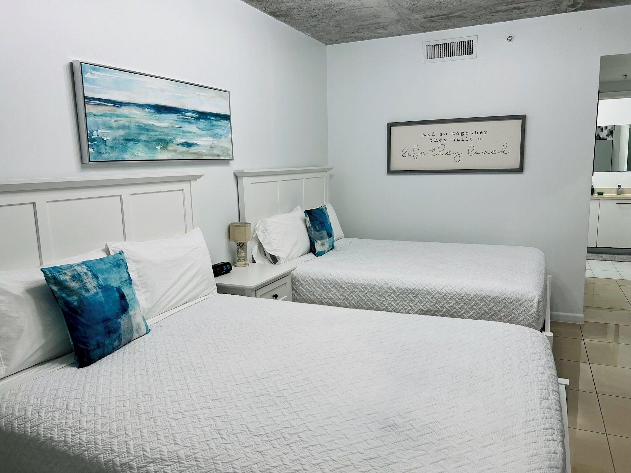 Habitación de hotel con dos camas, una mesita de noche, una lámpara y un cuadro de un océano azul en la pared.