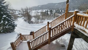 A wooden staircase leading to a deck with snow-covered railings, overlooking a snowy landscape with trees and buildings. - Large mountain home, incredible views, hot tub, game room, minutes from Jackson! (Jackson)