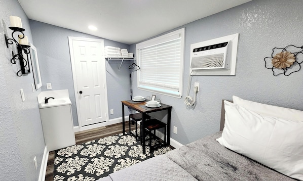 A compact room with a bed, a small table with a sink, a door, and a window with blinds.