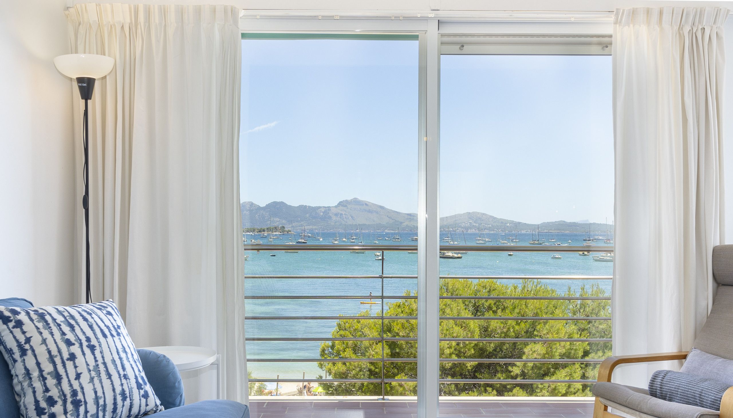 A room with a large sliding glass door leading to a balcony with a view of the sea and boats.