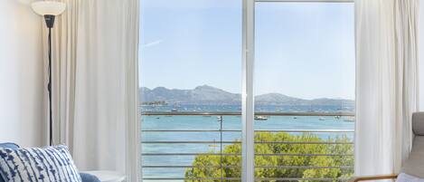 A room with a large sliding glass door leading to a balcony with a view of the sea and boats.