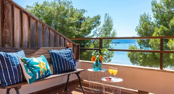 A terrace with a wooden bench, a small table with drinks, and a view of the sea and trees.