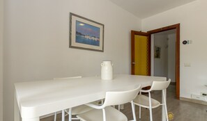 Un coin repas avec une table et des chaises blanches, un tableau encadré au mur et une porte dotée d’un cadre jaune.