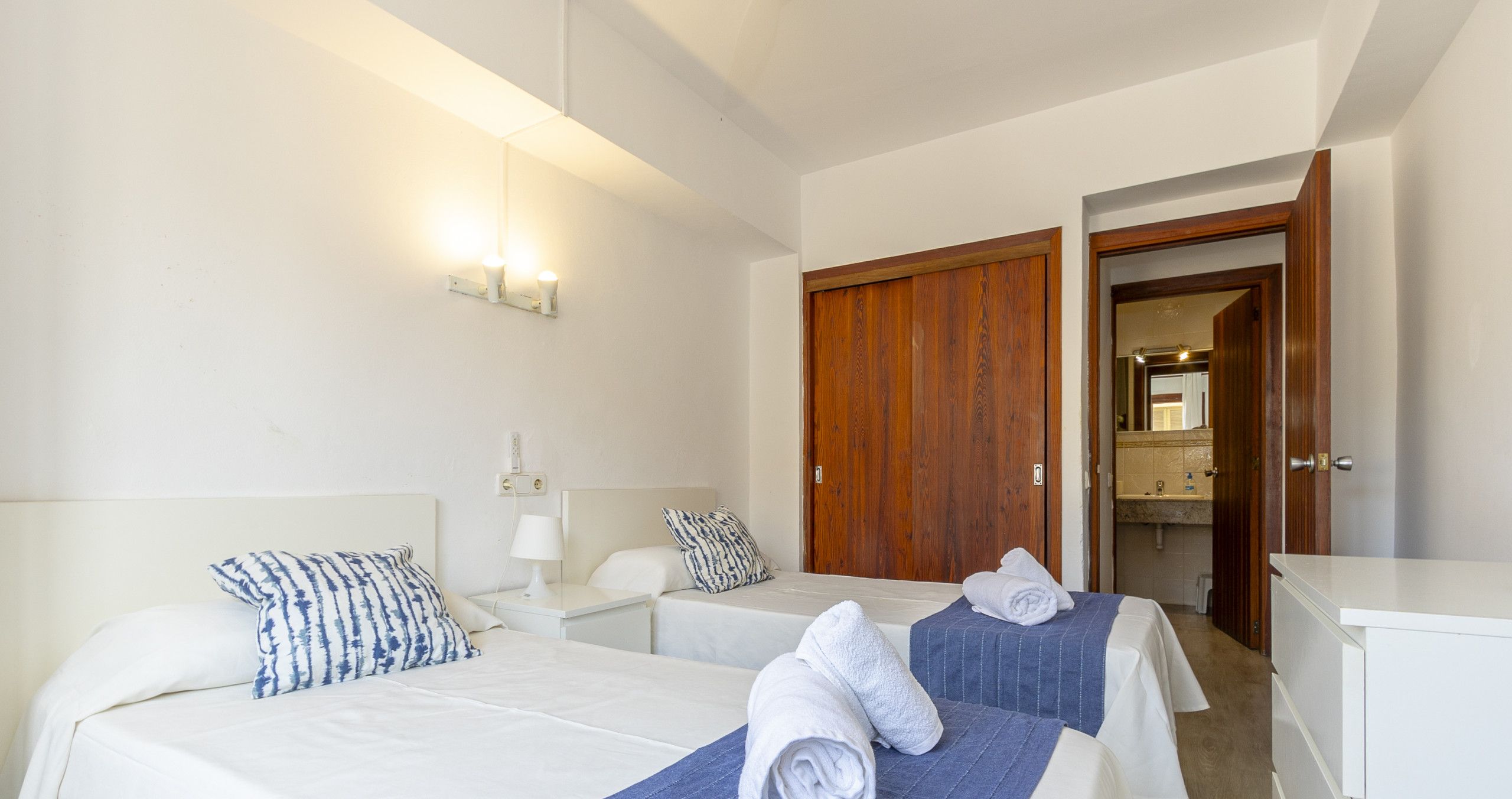 Een hotelkamer met twee bedden, een houten garderobe, een wit bureau en een badkamer zichtbaar door een open deur.