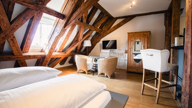 Ein Schlafzimmer mit schrägen Wänden, Holzbalken, einem Bett, einem Schreibtisch, einem Stuhl und einem Kleiderschrank.