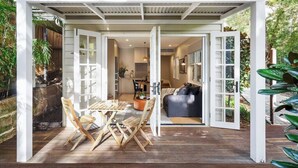 A covered outdoor dining area with wooden furniture and a view into a modern interior space. - Beautiful Balmain East Cottage (Balmain East)