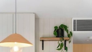 A modern kitchen with white cabinets, a hanging pendant light, a wooden shelf with plants and bowls, and a countertop with wine glasses and a bottle. - Beautiful Balmain East Cottage (Balmain East)