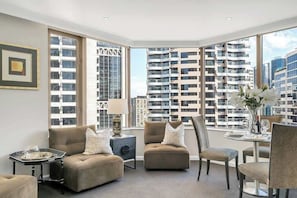 Una sala de estar moderna con sofá, sillón y mesa de comedor, ventanas amplias con vista a la ciudad y un cuadro en la pared.