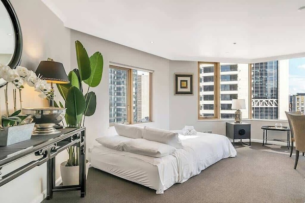 Una habitación de hotel moderna con una cama grande, un escritorio con lámpara, una planta en maceta y ventanas amplias que ofrecen vistas a la ciudad.