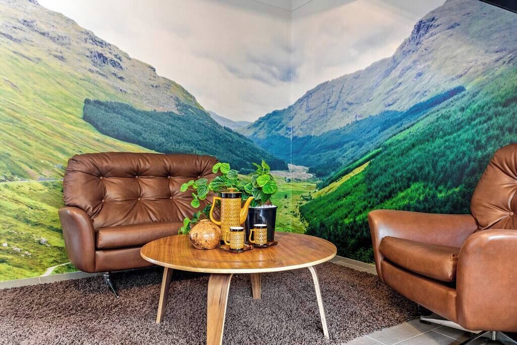 A room with a mountain landscape mural, two leather armchairs, a round wooden table with a plant and decorative items.