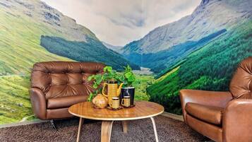A room with a mountain landscape mural, two leather armchairs, a round wooden table with a plant and decorative items.