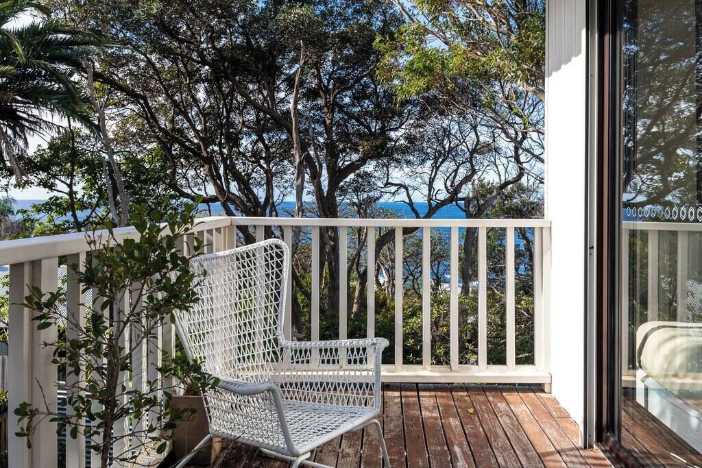 A balcony with a white metal chair and a wooden deck, overlooking a coastal area with trees and a view of the sea.