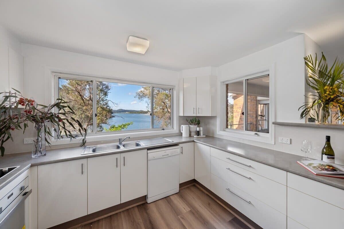 A modern kitchen with white cabinets, a dishwasher, and a large window offering a view of a lake and trees.
