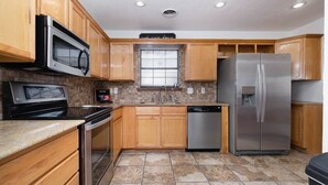 Una cocina con armarios de madera, electrodomésticos de acero inoxidable, una ventana con persianas y piso de baldosas.
