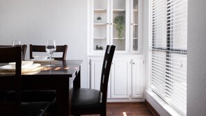 Un comedor con una mesa de madera dispuesta para dos personas, armarios blancos y una ventana con persianas.