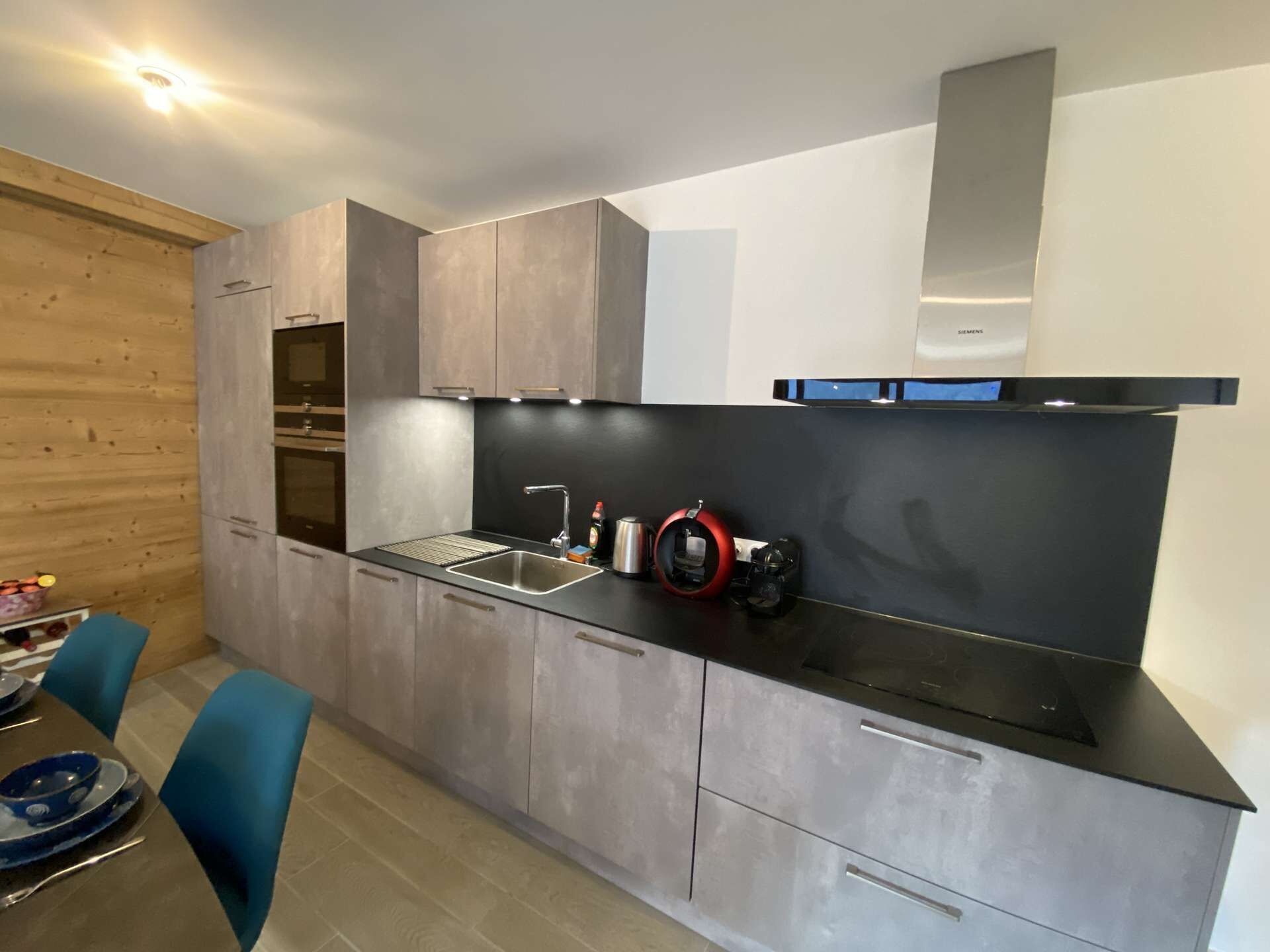 A modern kitchen with grey cabinets, a black countertop, and a Siemens range hood. There is a sink with a faucet and a red coffee machine. Blue chairs are visible at a dining table.