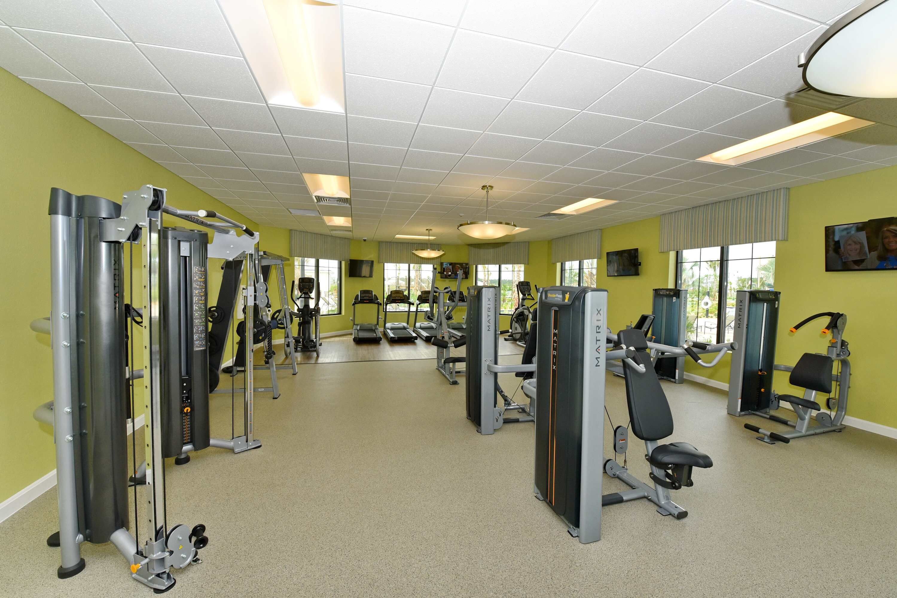 A gym with various exercise machines, large windows, and a yellow wall.