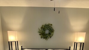 Cama bem arrumada com colcha branca, duas luminárias de cabeceira, travesseiro verde e manta xadrez.
