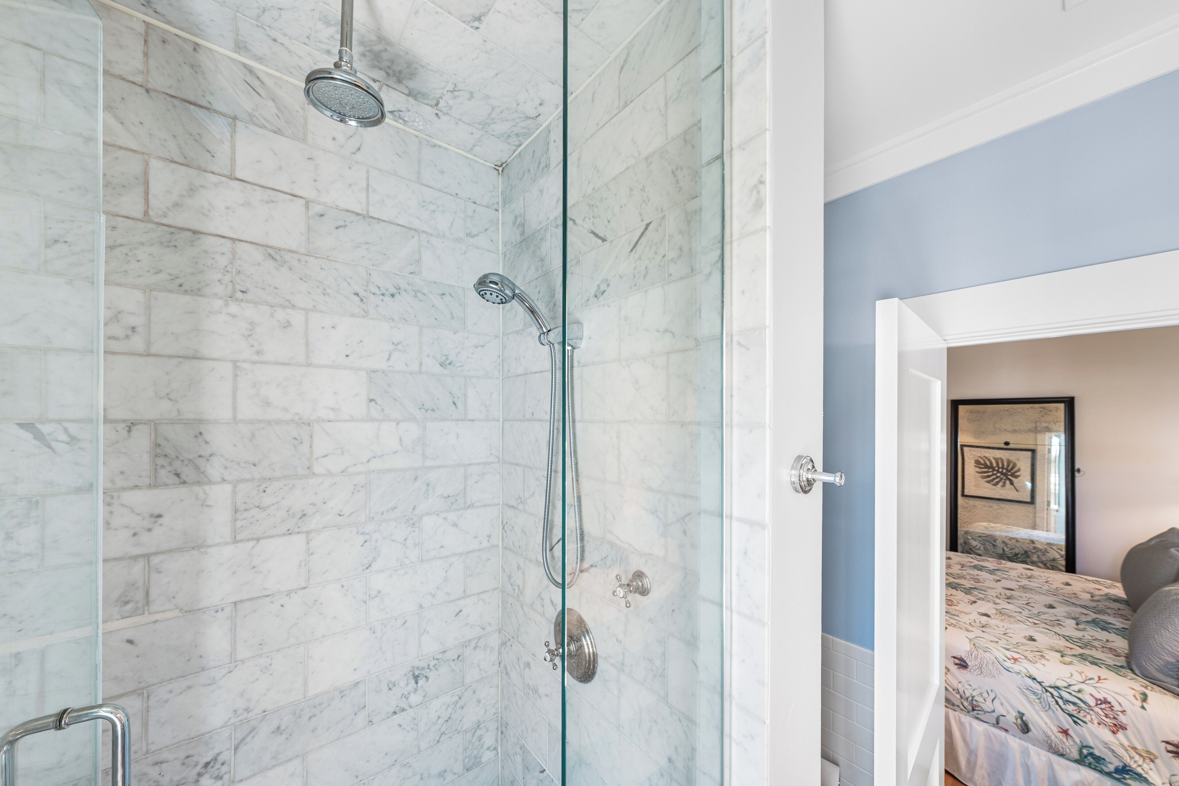 Une salle de bain dotée d’une douche à parois de verre, de carreaux de marbre et d’un lit recouvert d’une couvre-lit à motifs floraux, visible à travers une porte ouverte.