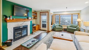 Una sala de estar con chimenea, televisor, sofá verde y vistas amplias a través de grandes ventanales.