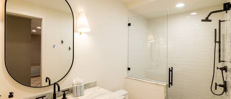 A modern bathroom with a large mirror, a sink with a marble countertop, a toilet, and a glass-enclosed shower area.