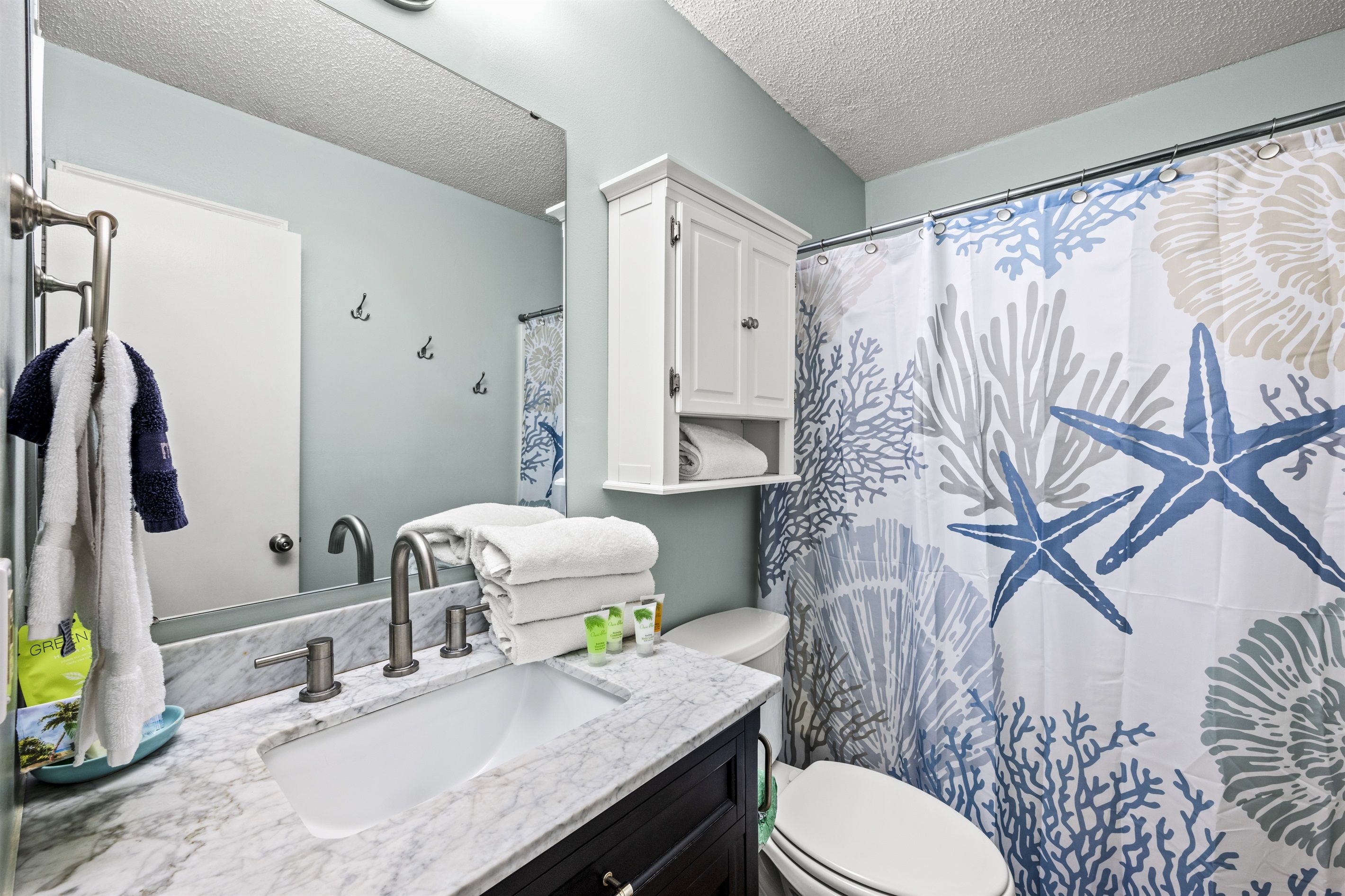 A bathroom with a marble sink, white cabinets, a white toilet, and a shower curtain with a starfish and coral pattern.