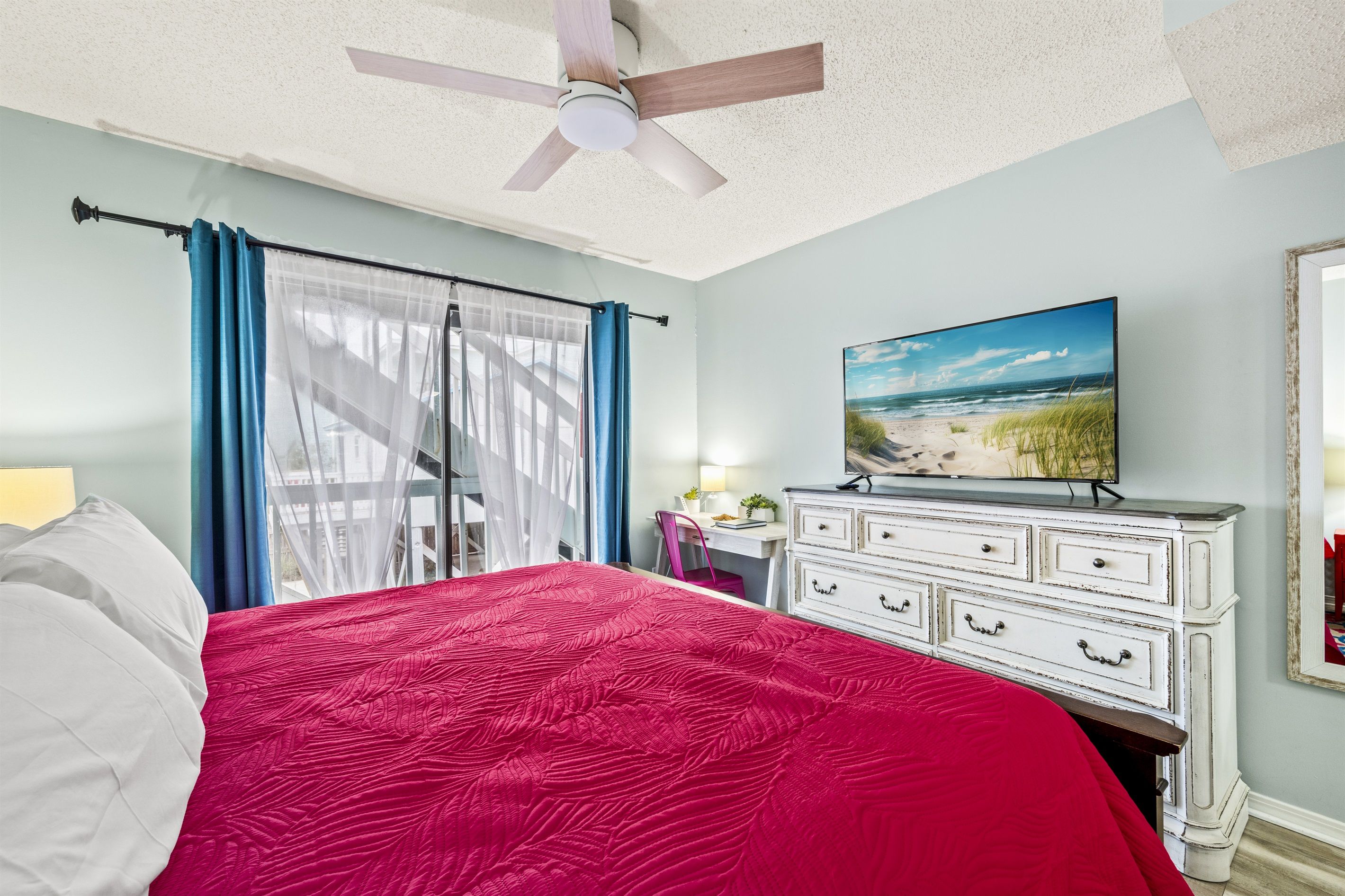 Une chambre à coucher avec un lit, un ventilateur de plafond, une commode, un tableau et une fenêtre avec des rideaux.