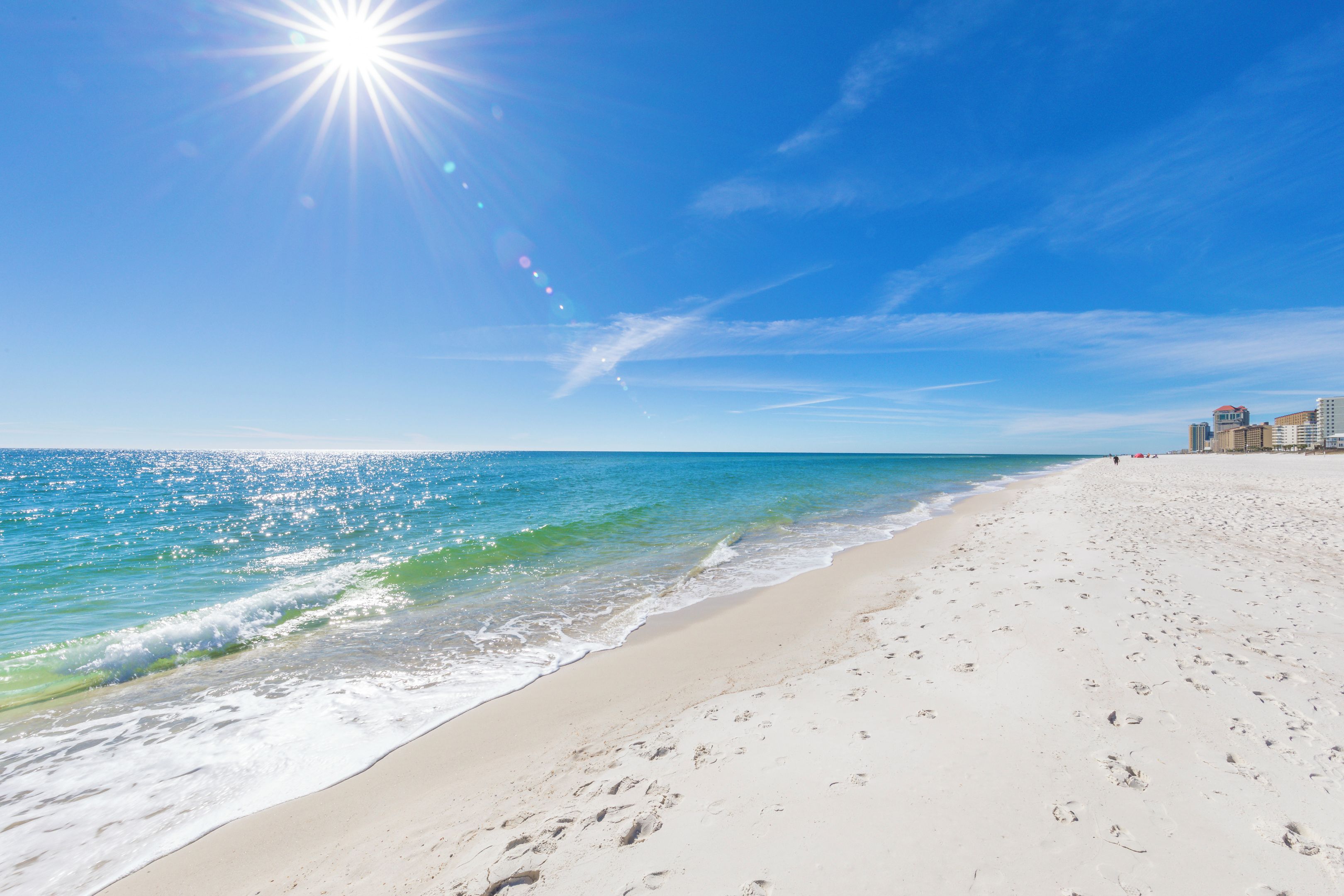 Ein Strand mit Fußabdrücken im Sand, ein klarer Himmel und Gebäude im Hintergrund.