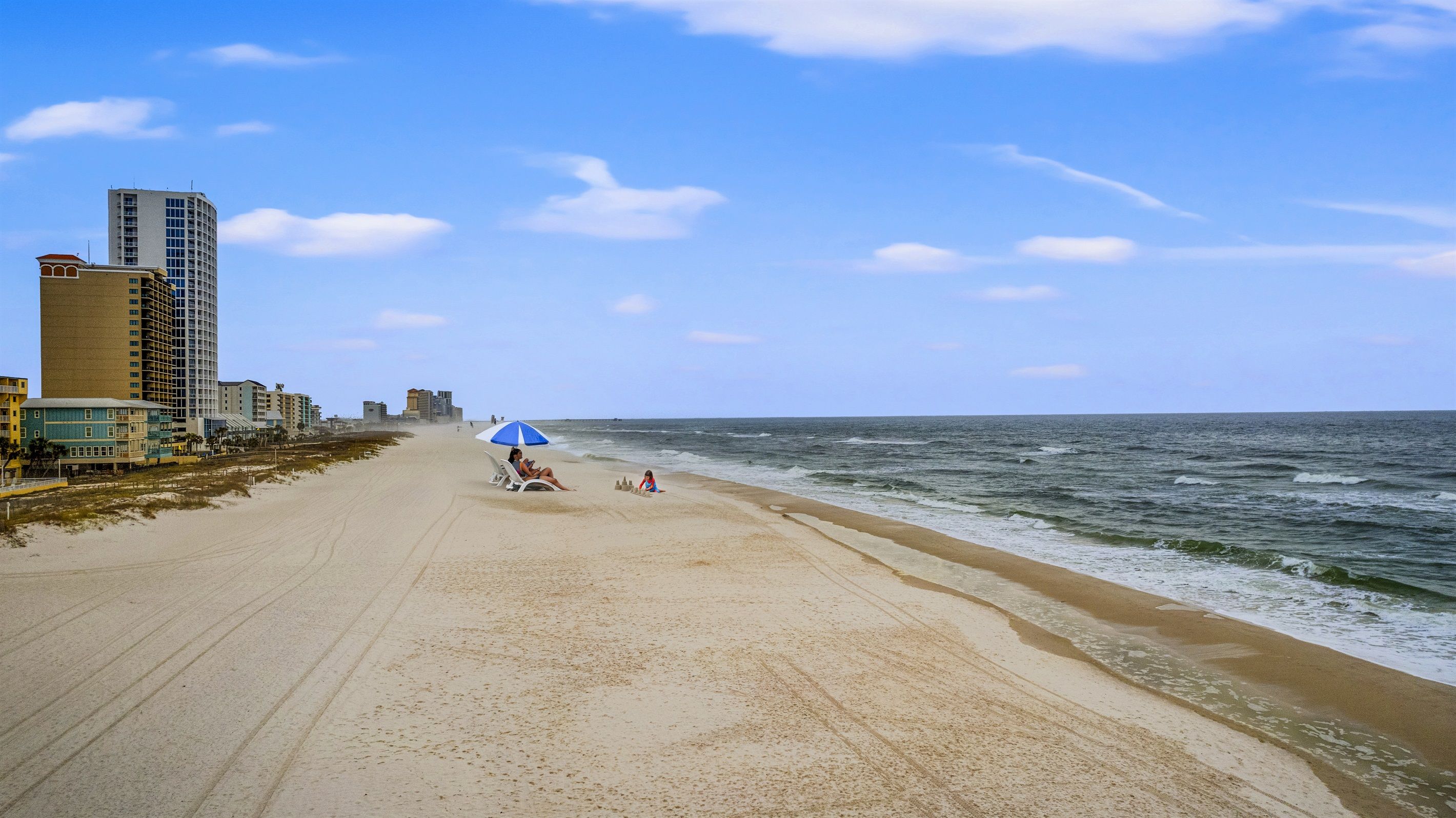 Una amplia playa de arena con cielo despejado, algunos edificios al fondo y una persona jugando en la arena.