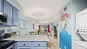 Une cuisine moderne avec un îlot central, un four à micro-ondes et un évier. On y trouve un coin repas avec une table et des chaises, ainsi qu’un salon avec un canapé et une table basse. Les murs sont peints en bleu clair et en blanc, et un panneau « Bienvenue » est accroché au mur.