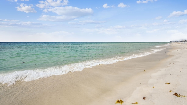 A beach with clear turquoise water, a sandy shore, and a few buildings in the distance.