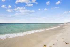 Une plage à l’eau turquoise et limpide, un rivage sablonneux et quelques bâtiments visibles au loin.